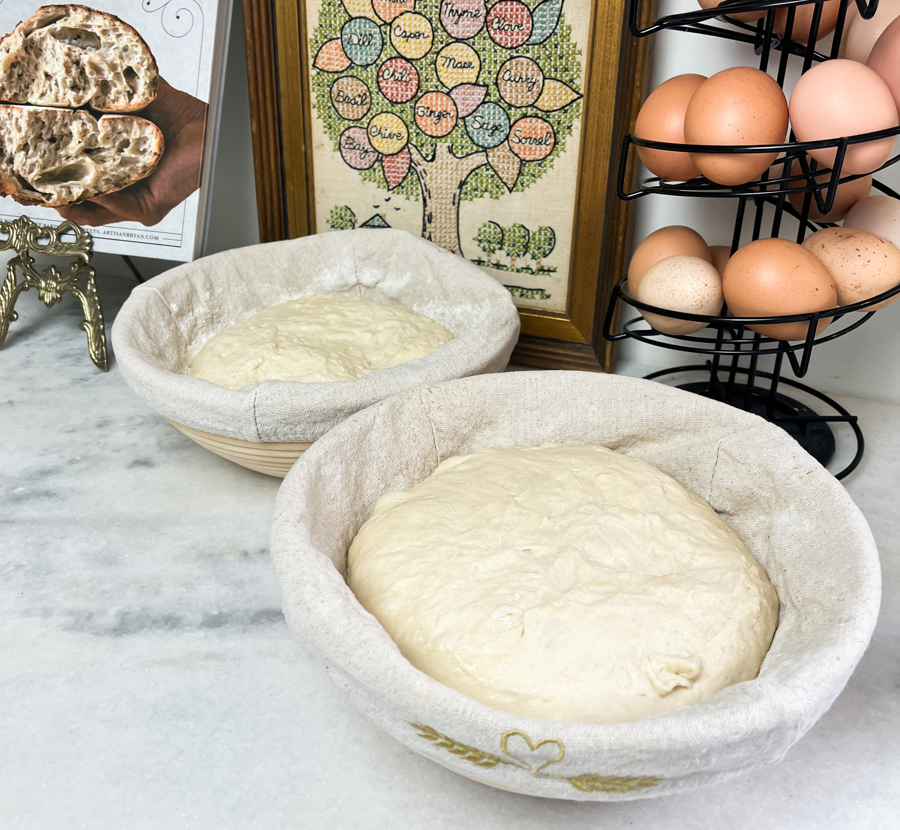 two cloth lined bowls with dough inside sitting on a marble countertop with brown eggs and a picture